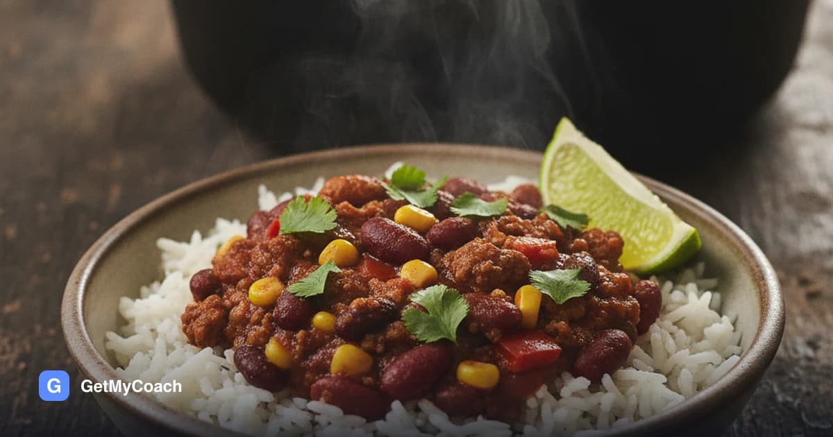 A deep bowl of dark red chili with kidney beans, black beans, sweetcorn and diced pepper over white rice, topped with chopped coriander and a wedge of lime.