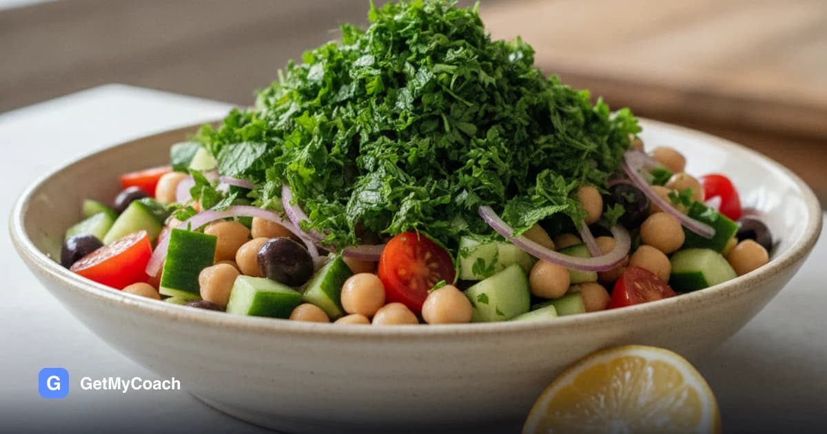 A wide bowl of chickpeas with diced cucumber, halved cherry tomatoes, black olives and thin red onion, under a thick layer of chopped parsley and mint, with a lemon half beside it.