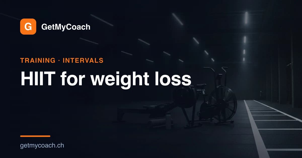 A rowing machine and an air bike side by side in a dark floodlit training hall, a marked sprint lane running past them — cover image for the GetMyCoach guide to HIIT for weight loss.