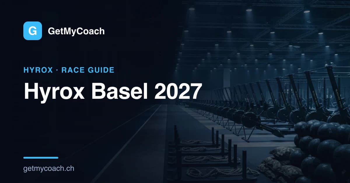 The floodlit floor of a large indoor exhibition hall set up for an indoor fitness race, with a rig of sleds and rowing machines waiting in the foreground — cover image for the GetMyCoach guide to Hyrox Basel 2027.