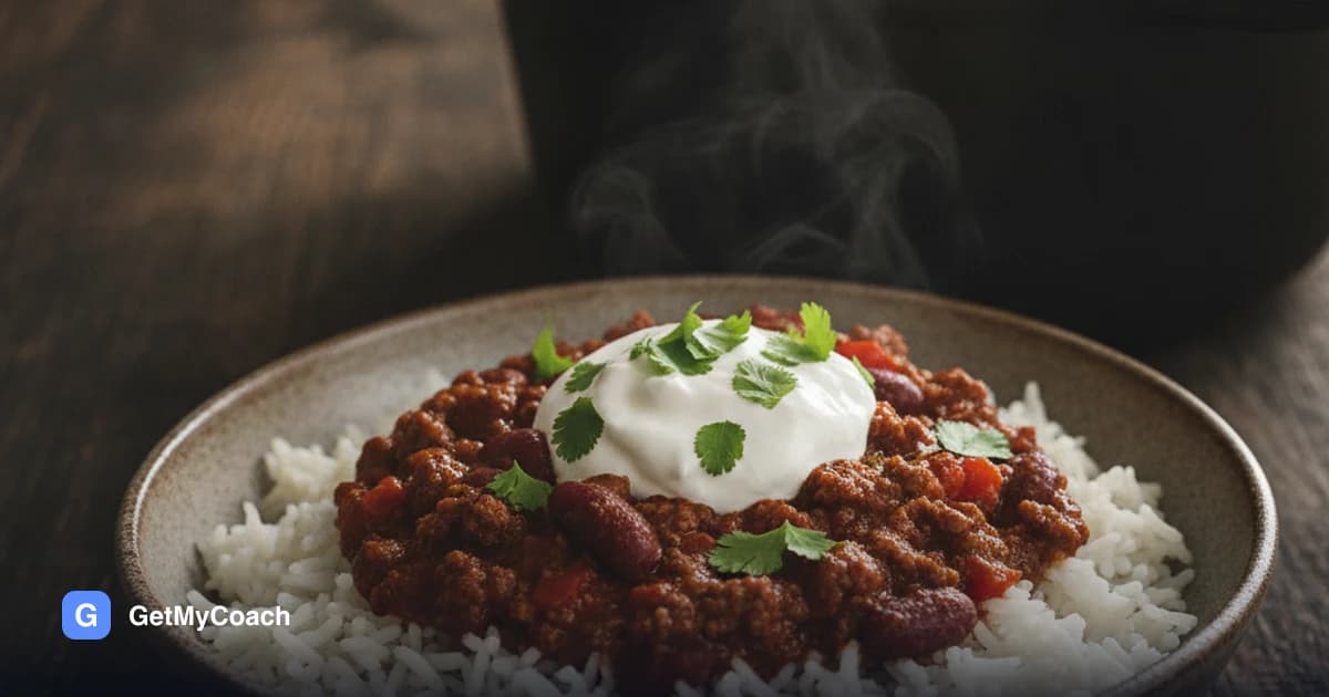 A deep bowl of dark red chili con carne over white rice, topped with a thick spoon of skyr and chopped coriander, with a second portion and a cast-iron pot behind it.